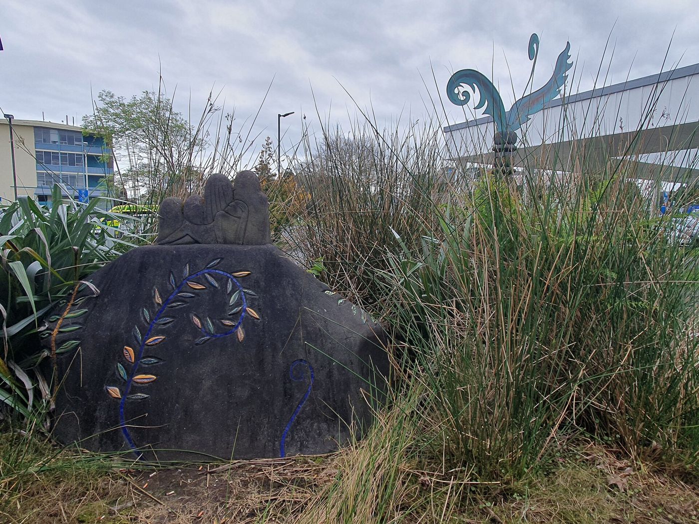 A carved dark stone with painted leaves and a metal sculpture on a pole stand among grasses in the Waitākere Hospital sculpture garden.